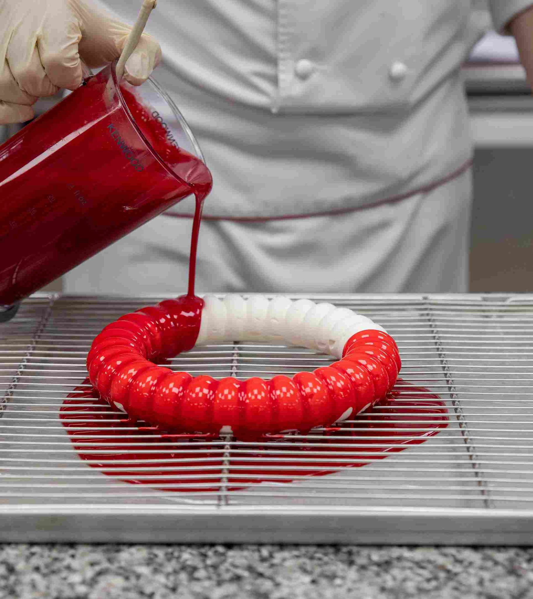 A chef pouring red liquid onto a circular mold with red and white coloring on a metal rack.