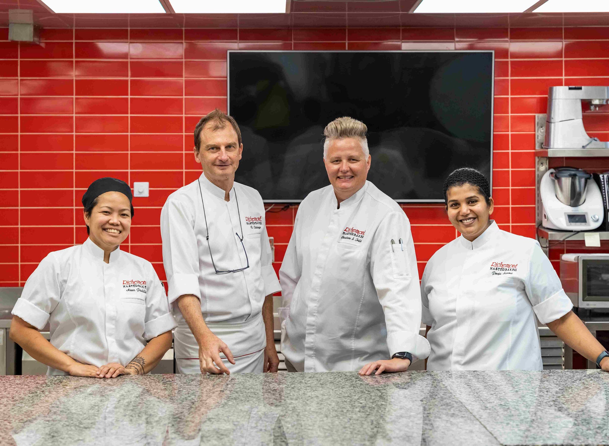 Four chefs in white Richemont Master baker jackets stand behind a granite counter in a kitchen with red tiled walls.